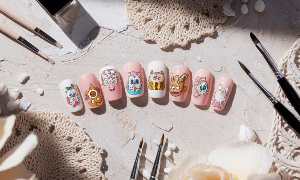 A set of artificial nails with cartoon designs including a strawberry, donut, face, ice cream, sneaker, bee, macaron, and hot dog. They are placed on a textured surface surrounded by brushes, crochet pieces, and decorative shells.