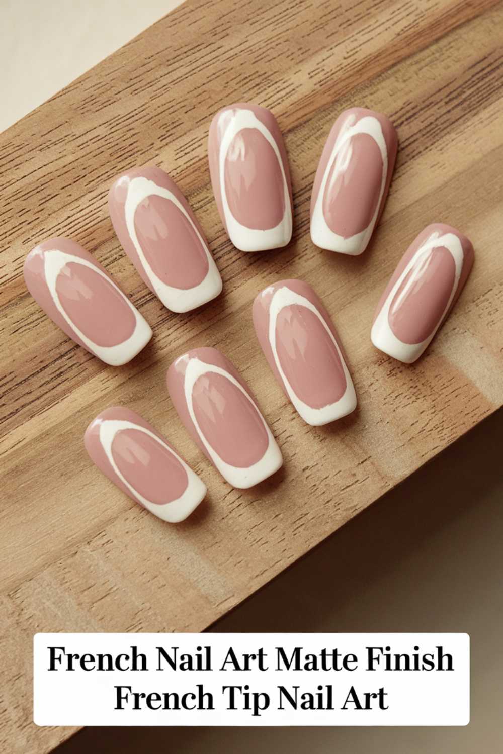 A set of press-on nails arranged on a wooden surface. The nails have a matte finish with a pink base and white French tips featuring an artistic oval design. A label at the bottom reads, French Nail Art Matte Finish, French Tip Nail Art.