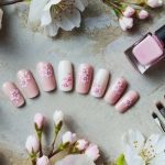 Pink floral nail art is displayed with a matching bottle of nail polish and brushes. White and pink cherry blossoms surround the scene on a textured surface.