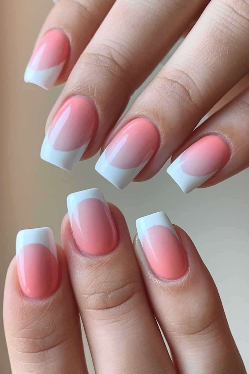 Close-up of hands with manicured nails showcasing simple nail art in a pink and white gradient design. The nails are short and square with a glossy finish, revealing a subtle transition from peachy-pink to white tips.