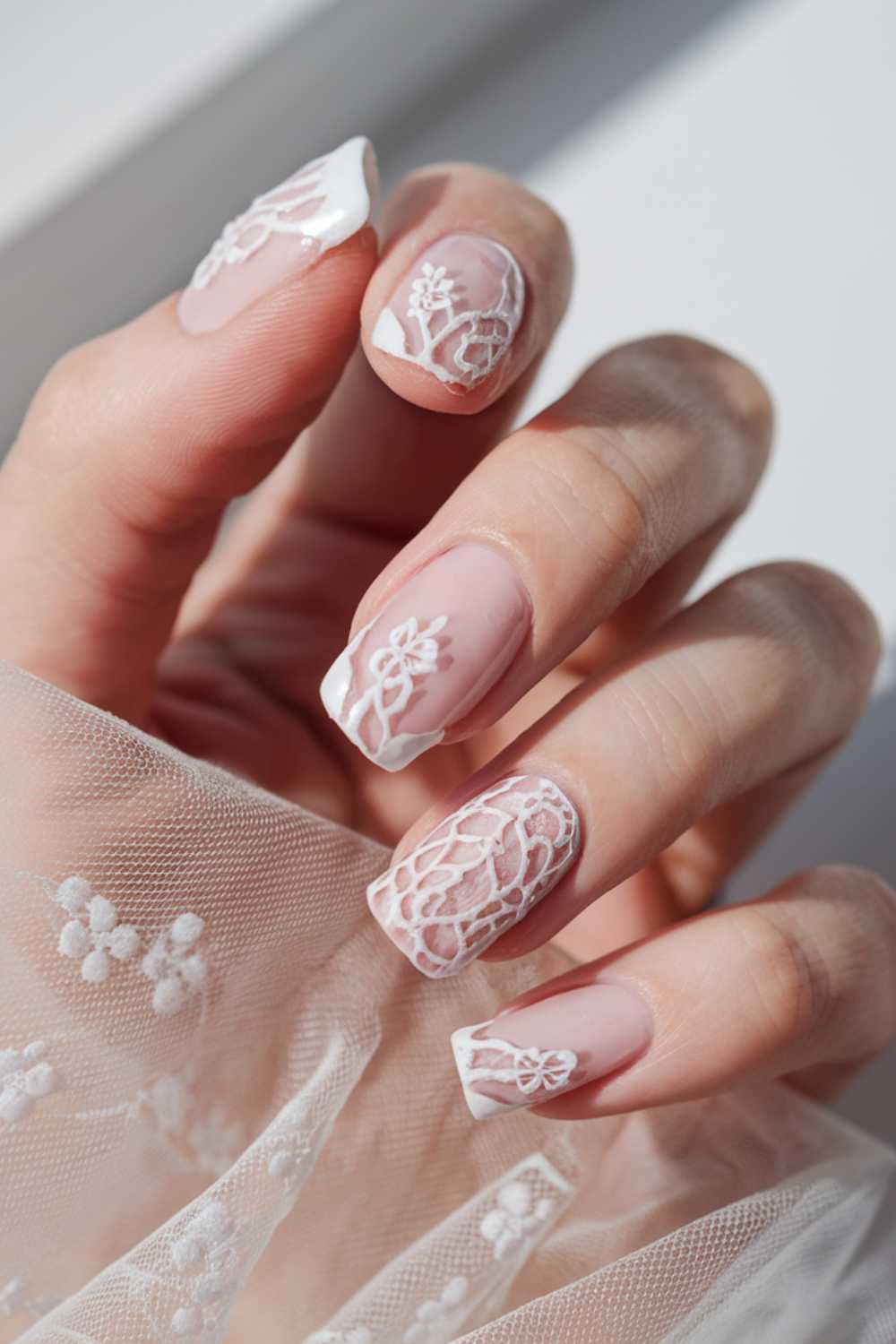 A hand with elegantly manicured nails featuring intricate lace-like white patterns over a soft pink base. The fingers hold a translucent fabric with floral embroidery, complementing the delicate nail art.
