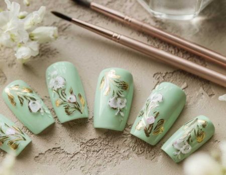 A set of mint green artificial nails featuring white flowers and gold leaf designs, arranged beside two nail art brushes and surrounded by delicate white flowers, perfect for showcasing elegant green nail art.