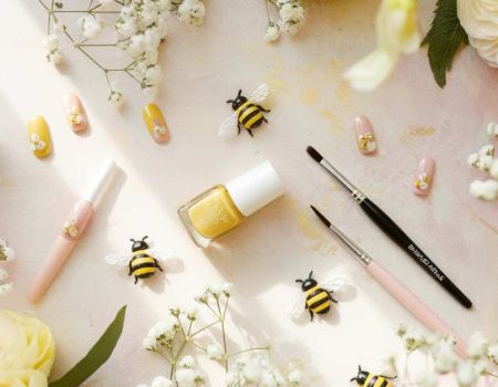 A flat lay of yellow nail polish, painted bee-themed fake nails, two brushes, small decorative bees, and white flowers on a soft, pastel-colored background. Surrounding are large cream and pink flowers.
