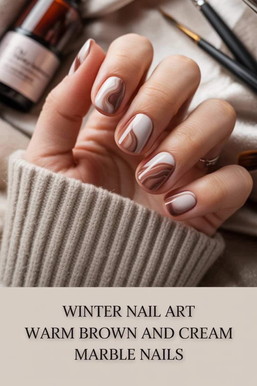 A hand with short nails features brown and cream marble nail art. The person wears a gray knit sweater, with nail polish bottles and brushes in the background. Text at the bottom reads: Winter Nail Art Warm Brown and Cream Marble Nails.